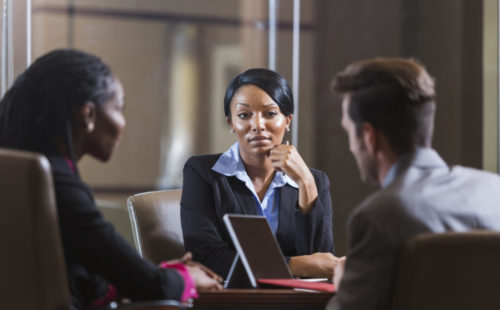 A group of three multi-ethnic businesspeople in a meeting in an office boardroom.  They are sitting, and there is a digital tablet on the table.  The focus is on the young African American woman with her hand on her chin, wearing a black suit.